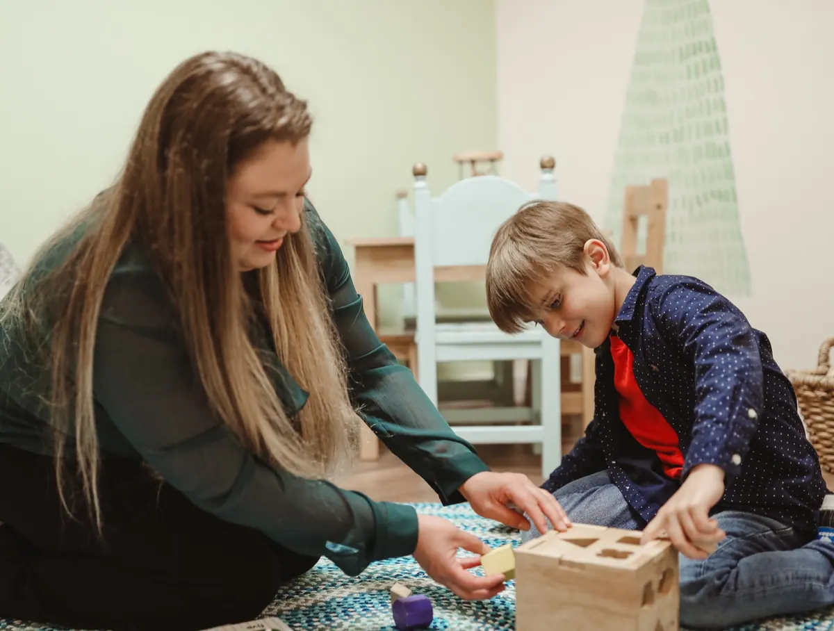 A therapist and a young boy sit on the floor sorting shapes together.