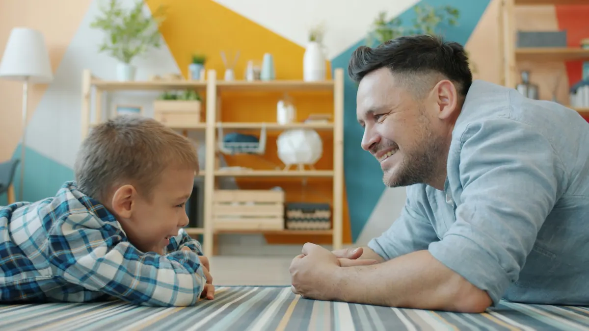 A father and son lying on the living room floor at home, smiling at each other