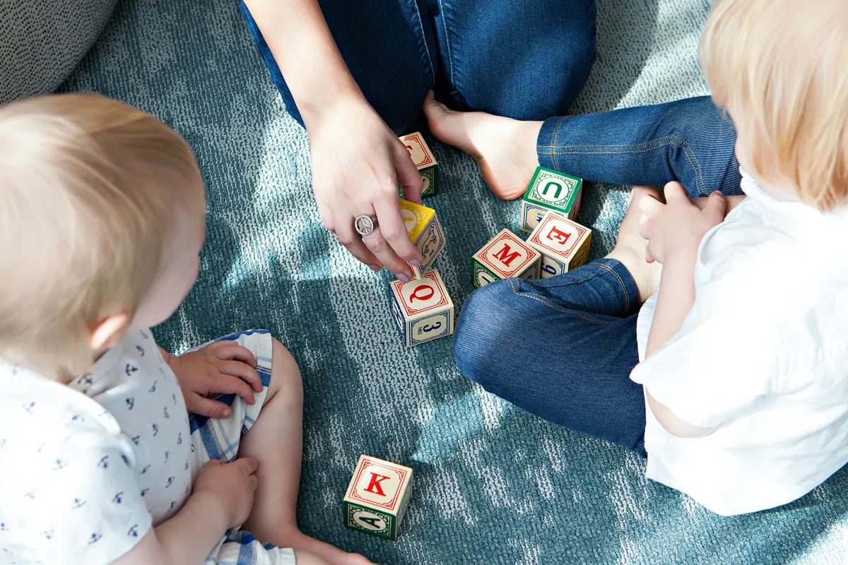 A caregiver playing with alphabet blocks with two children on the living room floor at home